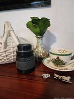 Photo showing white ceramic basket, camera lens, metallic vase with green floral arrangement, Christmas teacup and saucer partially visible, and two pins in front.