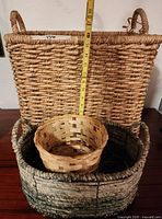 Three woven baskets displayed stacked on a wooden surface against a white wall. A measuring tape shows the height of the largest basket at about 15 inches tall.