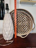 Photo of a tall contemporary modern white ceramic lamp with geometric diamond pattern base next to a large oval woven basket with checkerboard pattern, showing size reference with a ruler.