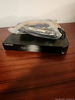 Samsung Blu-ray player on wooden surface with a plastic bag containing remote and cords on top.
