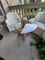 Front angle showing the round glass top and bamboo base side table placed between two cream upholstered chairs outdoors.