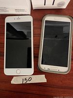 Front view of iPhone 6 Plus next to Samsung smartphone with protective case, both powered off, placed on wooden surface with handwritten '150' tag.