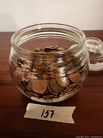 Clear glass jar filled with copper pennies, jar placed on wooden surface with lid off to the side.