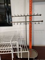 Metal jewelry rack with two horizontal bars and multiple hooks, bronze finish, shown against wall with measuring tape for size reference.