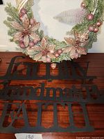 Photo of a large decorative wreath with pink poinsettias, green foliage, and pink ornaments, placed behind three black metal word signs 'Family', 'Grandmas place', and 'Faith'.