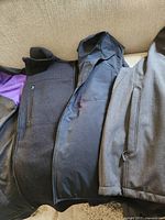 Three men's jackets laid side by side on a beige couch, showing black fleece vest, black Rockies jacket, and gray coat.