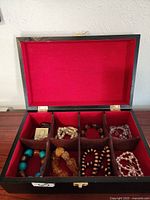 Open black jewelry box with red fabric interior showing six compartments filled with assorted bracelets made of various beads and materials.