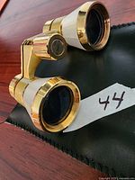 Close-up photo of gold-tone petite opera binoculars placed on black carrying bag, showing front lenses and adjustment knob.