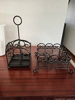 Photo of two black metal wire holders: a square napkin holder with a tall round handle and a rectangular silverware holder with curled feet and scrollwork around sides.