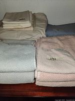 Multiple folded single bed sheet sets in beige and blue colors alongside textured pastel pink and light blue towels stacked on wooden surface.