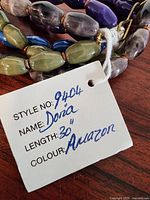 Close-up photo of various ceramic beads in purple, green, blue, and gray tones, showing their glossy finish and shape. Attached white tag with handwritten style number 9404, name Dora, length 30", color Amazon.