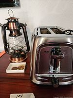 Photo showing Oster 2-slice toaster, vintage-style battery-operated lantern, and a small dish holding coins or tokens on a wooden table.