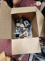 Open cardboard box showing various used hand tools and a tape measure inside, placed on a red carpet.