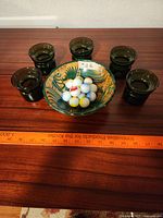 Top view of ceramic bowl filled with marbles surrounded by six green glass candle holders on wooden surface with measuring ruler for scale.