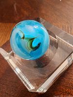 Close-up photo of a vintage glass marble with blue, white, and green swirls, displayed on a clear square base.