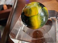 Front angle of a vintage glass marble showing distinctive yellow, green, and black swirls placed on a clear acrylic square stand.
