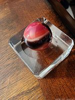 Photo showing close-up of the red, white, and black swirl glass marble resting on a clear acrylic square stand on a brown wood surface.
