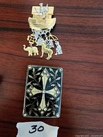 Photo of two vintage accessory pins on a wooden surface: a Noah's Ark pin with animal charms and a cross pin with decorative accents