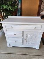 Front view of the vintage white side table dresser showing three drawers on the left and cabinet door with decorative embossed metal panel on the right.