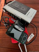 Sanyo cassette tape recorder with clear cassette door and side playback controls, alongside LG slide mobile phone with charger on wooden surface.