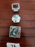 Three small vintage pill boxes arranged vertically on a wood surface: square abalone shell sterling silver box, round mother-of-pearl shell sterling silver box, square pewter box with reddish-brown stone top.