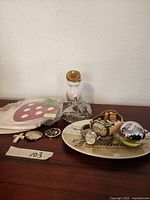 Wide view of lot items including watches, pocket watches, perfume bottle, note pads, and decorative plate on a wooden surface