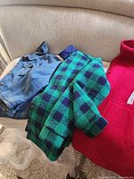 Three women's outerwear items displayed on a couch: a red coat, a blue denim jacket, and a green and navy checkered coat.