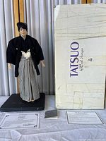 Full view of the traditional Japanese doll next to the original Tatsuo branded box, the doll wearing a black kimono and striped hakama on a black rectangular wooden base.
