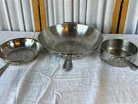 Three All-Clad stainless steel cookware pieces: frying pan (left), wok (center), saucepan (right) laid on white cloth with some wear visible.