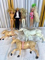 Four Barbie dolls standing against a white backdrop, hair disheveled, one male doll wearing tuxedo, two female dolls partially nude, one female doll in colorful swimsuit. Three horses lying in front, two tan, one white, all with painted hooves and harnesses.