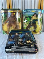 Three collectible dolls in original packaging: Barbie X-Files agent and two Ken dolls from Wizard of Oz in lion and scarecrow costumes. Packaging shows wear and aging.