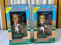 Front view of two boxed sports figurines, one of Cal Ripken Jr. in an Orioles uniform and one of Dave Justice in a Braves uniform, showing packaging wear and visible player faces inside clear windows.