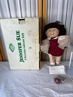 Jennifer Sue porcelain doll in red corduroy dress with lace collar and yarn hair standing beside original box and sealed accessory package.