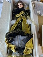Top-down view of porcelain doll in black and gold masquerade gown inside original box, showing full length of doll and gown details including gold mask and layered fabric.