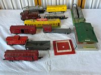 Overview of multiple vintage model trains and accessories arranged on a white surface showing red freight cars, yellow American Flyer boxcar, green building, and red platform accessory.
