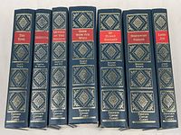Seven hardcover books standing upright showing spines with titles and authors, ornately designed in gold against dark blue leather and red title panels.