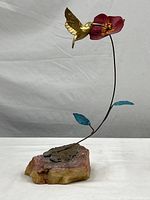 Full view of brass hummingbird feeding at the red metal flower atop the curved metal stem with blue leaves, attached to stone base.