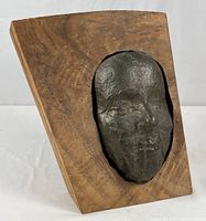 Front angled view of the bronze facial sculpture inset in a wooden plaque, showing the texture of the bronze and the natural wood grain.