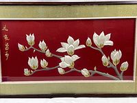 Close-up image of the embroidery showing white silk flower blossoms with gold thread outlining on red fabric background, with gold Chinese characters embroidered at the left side.