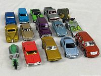Fifteen Hot Wheels scale model vehicles including cars, trucks, motorcycles, and a Jeep, all in various colors and styles arranged in three rows on a white surface.