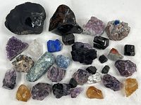 Top view of 27 unpolished mineral specimens of mixed colors including black, purple, blue, yellow, and clear crystals arranged on a white surface.