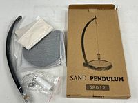 Photo of the sand pendulum components laid out alongside the box. Shows pendulum arm, base tray, pendulum weight, plastic tubing, and small hardware pieces.