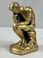 Front angled view of the brass sculpture showing the seated figure with hand on chin and textured base.