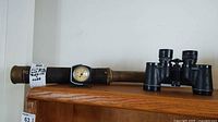 Antique maritime brass telescope with leather wrapping and an attached Tel-Tro marine thermometer, displayed on a wooden surface beside a pair of binoculars.