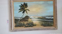Front view of the signed canvas print showing a beach with a leaning palm tree and waves in the background, framed in a distressed light wood frame.