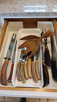 Drawer containing vintage carving utensils and wooden salad servers, showing variety of horn and antler handled knives and forks, and polished wooden salad server set.