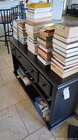 Full view of the black distressed console table filled with stacked books, showing the three drawers on top and two lower shelves including the wine rack.