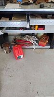 Overview of workbench lower shelves showing red gasoline container, coiled extension cords, small paint can, Black and Decker sander, sanding pad attachments, and workshop items.