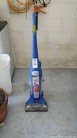 Full view of the blue Hoover floor cleaner standing upright on a concrete floor near a wall and some potted plants.