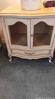 Full front view of one French Provincial nightstand by Drexel showing distressed cream finish, wire mesh front doors, single drawer with ornate handle, curved base with cabriole legs.